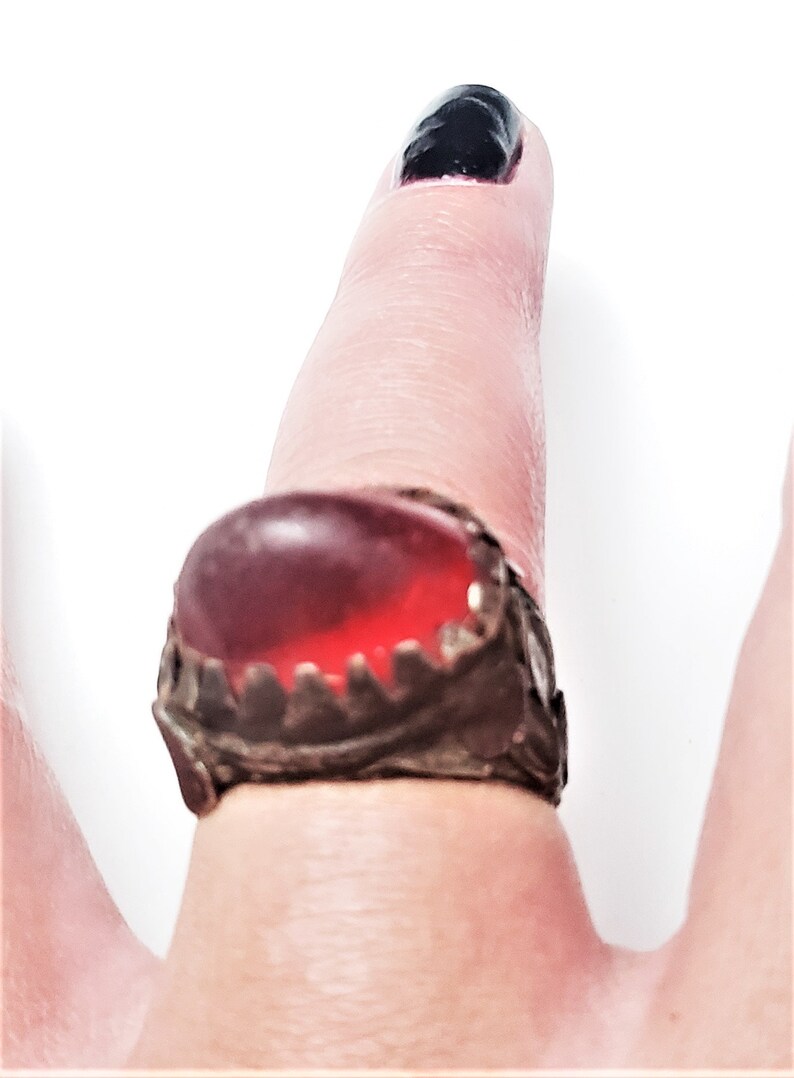 Puede incluir: Un anillo de plata con una piedra preciosa roja engastada en un engaste decorativo y dentado. El anillo se lleva en un dedo.