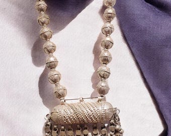 May include: A silver necklace with a pendant featuring a cylindrical charm with a tassel of small silver beads. The necklace is made of round silver beads with a textured surface.