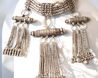 May include: A silver necklace with a choker style base and three dangling charms. The charms are made of silver beads and have a woven chain design.