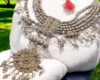 May include: An elaborate silver necklace with multiple strands of small beads and detailed pendant designs. The necklace features a large, ornate pendant with dangling elements and a red tassel. The necklace is displayed on a white surface.