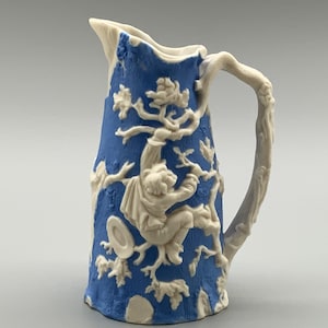 May include: A blue and white ceramic jug with a detailed relief design. The jug features a blue background with a white relief of a figure climbing a tree. The handle is shaped like a tree branch.
