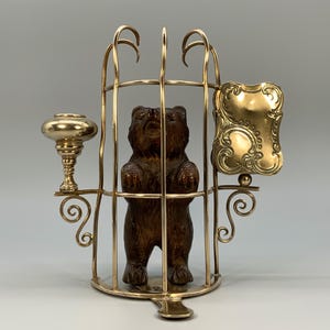 May include: A decorative gold-toned metal cage with a carved wooden bear inside. The cage has ornate scrollwork and a small, decorative gold-toned element on the side. A gold-toned candlestick holder is on the left.