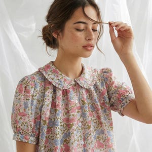 May include: A floral print blouse with a rounded collar. The short-sleeved top features a pattern of pink, blue, and green flowers on a cream background. The blouse has a button-down front and a ruffled collar.