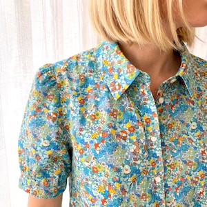 May include: A blue floral shirt with a button-down front and short, puffed sleeves. The pattern features a mix of blue, yellow, orange, and white flowers. The shirt has a classic collar and appears to be made of a light fabric.