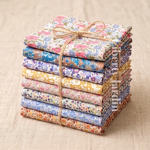 May include: A stack of ten folded fabric squares featuring various floral patterns in shades of pink, blue, yellow, purple, and orange. The fabric stack is tied together with a thin brown twine string. The text Liberty Jardin is visible.