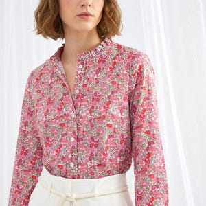 Peut inclure: Un chemisier rose à imprimé floral avec un col à volants et des manches longues. Le chemisier a une fermeture boutonnée sur le devant et est associé à un pantalon blanc. Le motif floral comprend des fleurs rouges, roses et violettes.