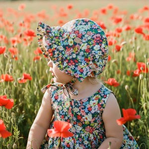 May include: A child wearing a floral print sun hat and dress in a field of red poppies. The hat has a ruffled brim and ties under the chin. The dress is sleeveless and also features a floral pattern. The background is filled with red poppy flowers.