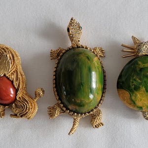 May include: Three vintage brooches: a poodle with a red stone, a turtle with a green stone, and a cat with a green and yellow stone. All are gold-toned metal with rhinestone accents.