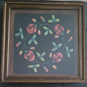 May include: A square framed artwork featuring a dark black background with a central circular wreath design. The wreath is composed of embroidered red and pink rose-like flowers connected by thin green stems with small green and pink buds.
