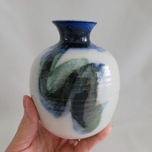 May include: A handmade ceramic vase with a white base and a dark blue rim. The vase features abstract brushstrokes in shades of blue and green. The vase is a decorative item.