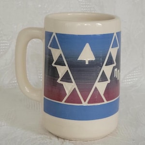 May include: A cream-colored ceramic mug featuring a horizontal band with a gradient from blue to dark purple. The band is decorated with a repeating pattern of white triangles and a single white pine tree silhouette. The mug has a sturdy handle.