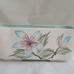 May include: Rectangular ceramic planter with a speckled, off-white finish. The planter features a hand-painted floral design with blue and pink flowers, green leaves, and a light green interior. A decorative piece for indoor or outdoor use.