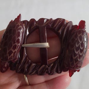May include: A vintage burgundy-colored belt buckle with a carved design. The buckle features a central circular opening with a silver-colored pin. The sides of the buckle have carved, textured details, and the overall design is ornate.
