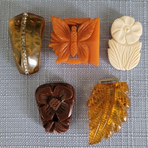 May include: A collection of five decorative dress clips and pins. The set includes a translucent amber piece with a rhinestone strip, an orange butterfly-shaped clip, a cream-colored floral piece, a dark brown flower, and a golden amber leaf-shaped clip.