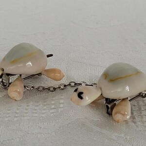 May include: Two turtle-shaped brooches crafted from polished off-white and pale blue seashells. Each turtle features a smooth shell, small head with black painted eyes, and four flipper-like appendages. The two brooches are connected by a silver chain.