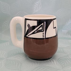 May include: A ceramic coffee mug with a white handle and a brown base. The upper portion of the mug is white with black geometric designs. The interior of the mug is off-white.