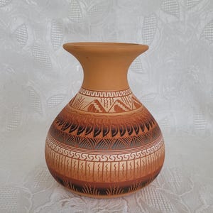 May include: A terracotta vase with a wide, flared rim and a rounded body. The surface features intricate hand-painted geometric patterns in dark brown and cream, including triangular motifs, repeating vertical lines, and stylized grass-like details.