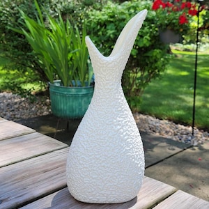 May include: A textured, off-white ceramic vase with a unique, elongated shape. The vase has a wide, rounded base that tapers to a narrow neck, then flares out into a distinctive, open top. The surface has a rough, pebbled texture.