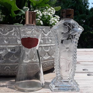 May include: Two vintage perfume bottles. One is clear with a red label and a copper-colored cap. The other is a clear, rectangular bottle with a floral design and a dark brown cap. Both bottles are on a wooden surface.