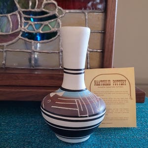 May include: A small, hand-painted Naatsilid Pottery vase with a white neck and a bulbous body. The body features a geometric design in brown, blue, and white, with black stripes. The vase is approximately 6 inches tall.