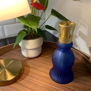 May include: A cobalt blue glass fragrance lamp with a ribbed, hourglass-shaped body and a gold-toned metal vented cap. The lamp features a smooth, matte finish and sits upright, showcasing a distinct vertical fluted texture along its base.