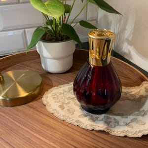 May include: A deep red, ribbed glass fragrance lamp featuring a shiny gold-toned metal crown top. The vessel has a rounded, bulbous base and sits on a small, oval-shaped fabric doily with a delicate, light-coloured floral embroidery pattern.