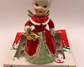 Może przedstawiać: Ceramiczna figurka anioła w czerwonym płaszczu z białym wykończeniem i jasnozielonej spódnicy, trzymająca złoty dzwonek. Anioł ma białe włosy z teksturowaną koroną i złote skrzydła. Figurka stoi na pudełku z napisem Christmas Tree Ornaments.