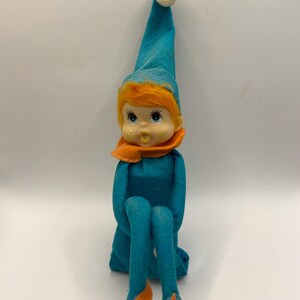 Peut inclure: Une poupée elfe vintage avec une tenue turquoise, comprenant un chapeau pointu avec un pompon blanc et des jambes turquoise assorties. La poupée a des cheveux roux, une écharpe orange et des chaussures orange. La poupée est assise.