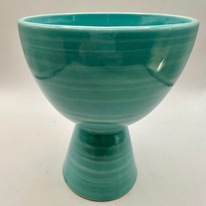 May include: A teal ceramic bowl with a pedestal base. The bowl has a wide, open top and a rounded shape, supported by a conical base. The surface has horizontal textured lines, and the colour is a vibrant teal.