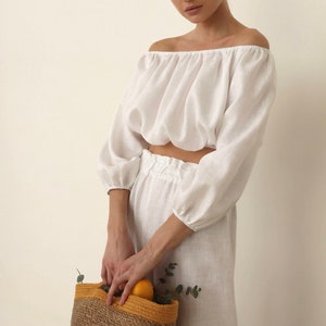 May include: A woman wearing a white off-the-shoulder linen top and matching pants holds a woven basket with a yellow trim. The basket contains oranges and eucalyptus leaves.