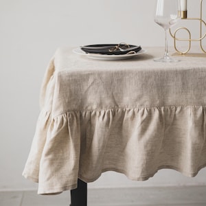 May include: A beige linen tablecloth with a ruffled edge covers a table set for a meal. A white plate holds a black plate, with silverware and a napkin. A wine glass and a gold candle holder with white candles are also on the table.