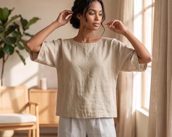 May include: A beige short-sleeved top with a horizontal seam across the midsection, paired with light gray trousers. The top features a wide, loose fit with elbow-length sleeves and a simple round neckline, crafted from a textured fabric.
