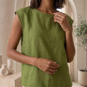 May include: A sleeveless olive green top with a round neckline and a straight hemline. The garment features a textured fabric finish with visible weave details, a relaxed fit, and a simple construction with wide shoulder coverage.
