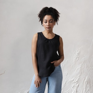 May include: A black sleeveless top with a gathered neckline. The top is made of a textured fabric and is paired with light blue jeans. The model is standing in front of a textured white wall.
