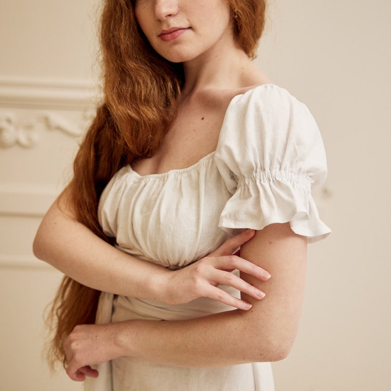 May include: A white linen dress with a square neckline and puffed sleeves. The dress is cinched at the waist with a matching fabric belt.