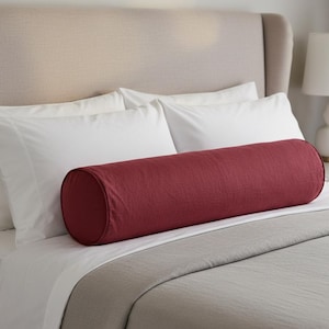 May include: A burgundy cylindrical bolster pillow rests on a bed with white sheets and a gray blanket. The pillow is made of a textured fabric and is positioned horizontally across the bed. The headboard is a light beige color.