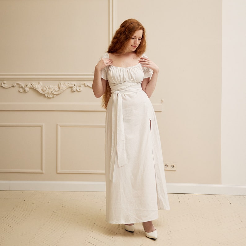May include: A white maxi dress with a square neckline, short sleeves, and a tie at the waist. The dress has a thigh-high slit on the right side.