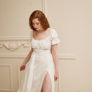 May include: A white linen dress with a square neckline, short sleeves, and a tie at the waist. The dress has a thigh-high slit on the left side.