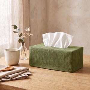 May include: A rectangular tissue box cover made of olive green fabric with a textured weave. The cover is placed over a standard tissue box, with white facial tissues protruding from the top opening. The fabric has visible stitching along the edges.