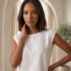 May include: A sleeveless white top featuring a round neckline and a straight-cut silhouette. The garment is constructed from a textured, woven fabric with a visible weave pattern, providing a clean and simple aesthetic for this basic wardrobe piece.