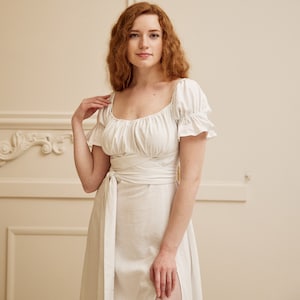 May include: A white linen dress with a square neckline and a wrap tie at the waist. The dress has short, puffed sleeves and a flowing skirt.