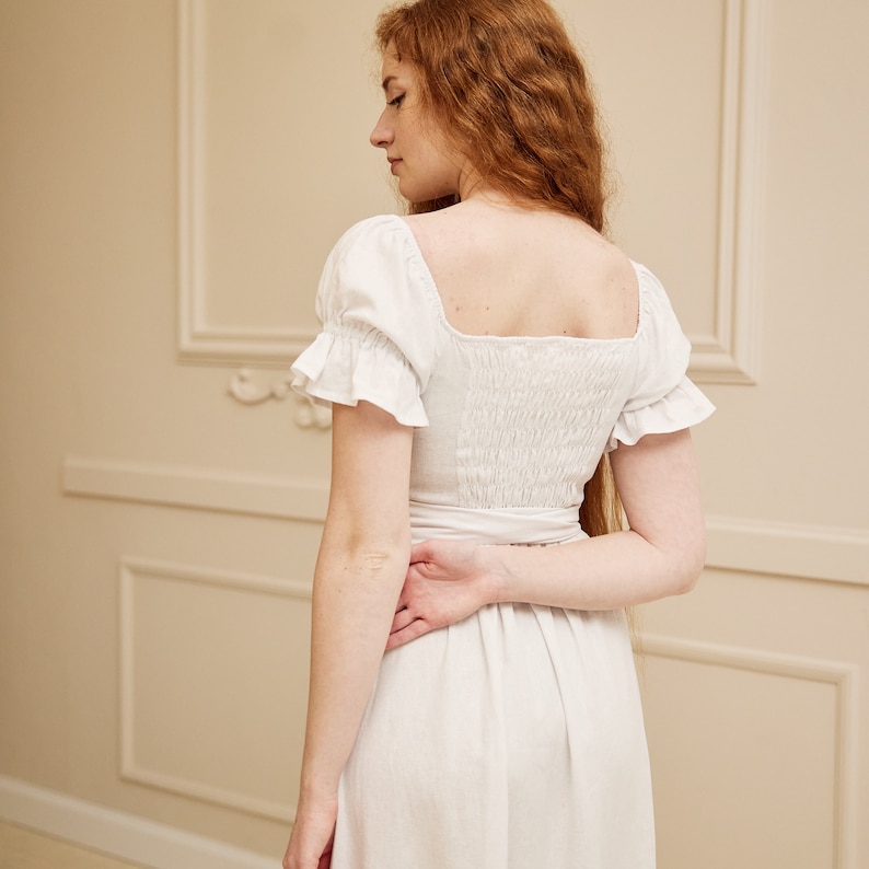 May include: A white linen dress with a square neckline and short puff sleeves. The dress has a fitted bodice and a flowing skirt.