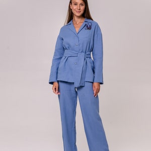 May include: A light blue linen pantsuit with a long-sleeved collared shirt and matching trousers. The shirt has a belt and a floral embellishment on the chest.