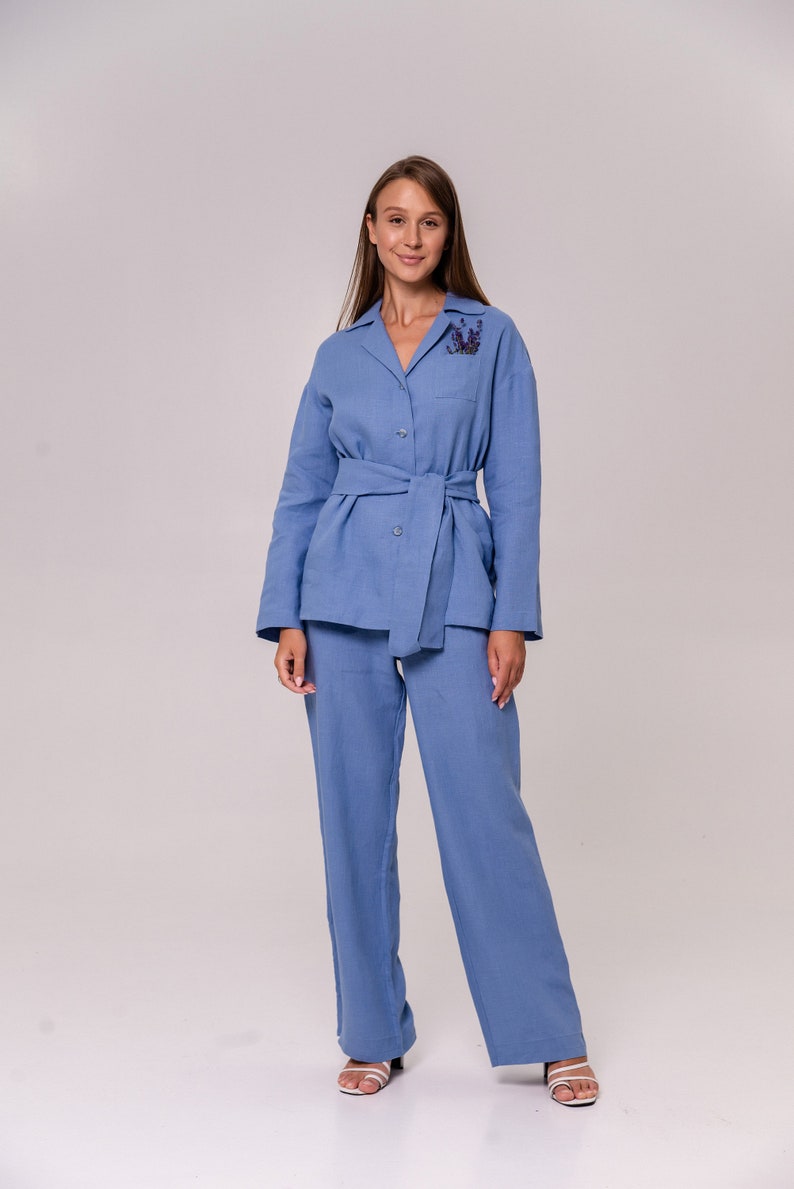 May include: A light blue linen pantsuit with a long-sleeved collared shirt and matching trousers. The shirt has a belt and a floral embellishment on the chest.