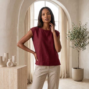 May include: A sleeveless, deep burgundy top with a round neckline and a straight hemline, paired with light beige trousers. The top features a textured fabric finish, while the trousers have a structured fit with a visible center seam detail.