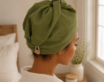 May include: An olive green hair towel wrap made of textured fabric, featuring a gathered design at the top and a secure closure at the nape of the neck consisting of a white elastic loop fastened around a round, light-colored wooden button.