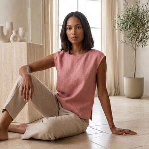 May include: A dusty rose sleeveless top with a round neckline and a straight hem featuring side slits, paired with light beige trousers. The top is constructed from a textured fabric, and the trousers have a relaxed fit with a visible weave pattern.