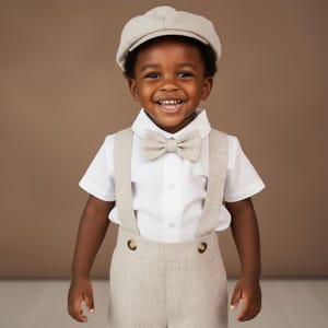 May include: A young child wearing a light beige newsboy cap, bow tie, suspenders, and shorts. The outfit includes a white button-down shirt. The child is smiling, with a brown background.