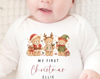 May include: A white long-sleeve bodysuit featuring a graphic of three teddy bears dressed in holiday attire, with the text "MY FIRST Christmas ELLIE 2026" printed below. The garment is paired with a solid rust-colored fabric headband with a bow.