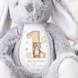Op de afbeelding: Grijze pluche konijn met een witte vlek op zijn borst. De vlek heeft een bruine beer die een ballon vasthoudt en een houten nummer &eacute;&eacute;n met gouden sterren. De tekst "Happy First Birthday William 02.04.2024" staat op de vlek.